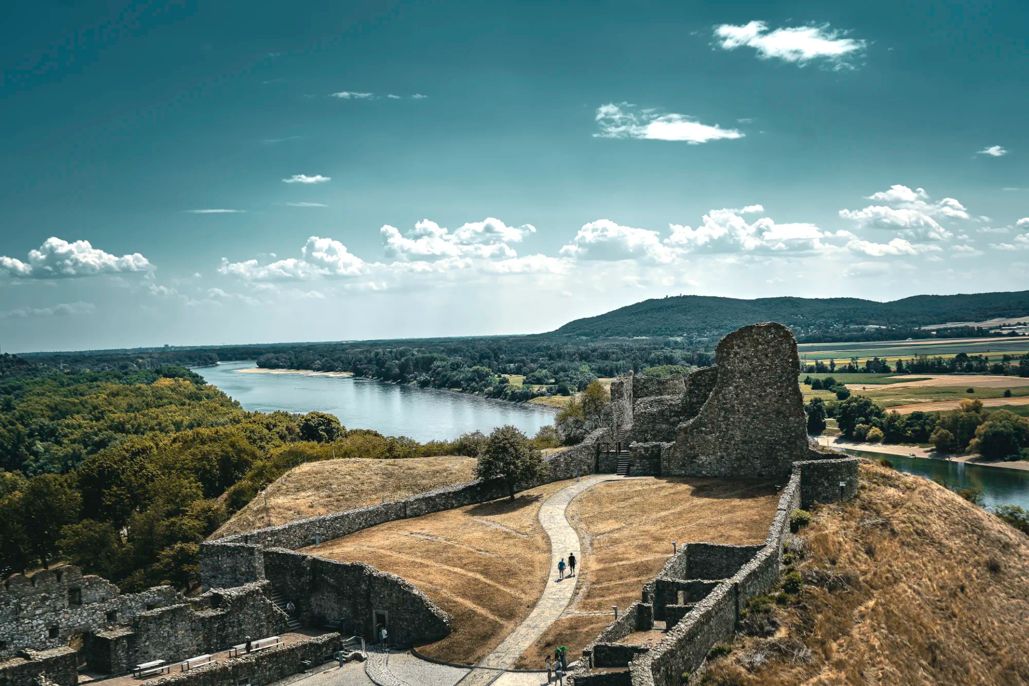 Devín Castle ruins above the confluence of the Morava and Danube, with fields and forest under a broad clouded sky.